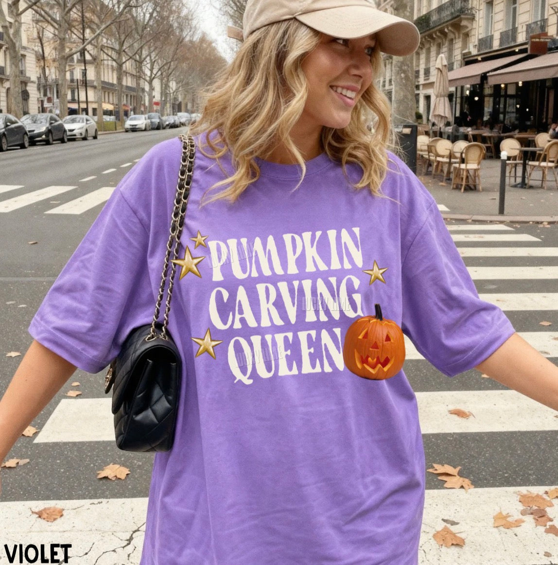 A woman wearing an oversized violet graphic T-shirt with the text 'PUMPKIN CARVING QUEEN' in white, decorated with gold stars and a jack-o'-lantern graphic. She is also wearing a beige cap and carrying a black shoulder bag, standing on a city crosswalk.