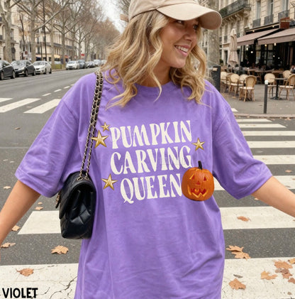 A woman wearing an oversized violet graphic T-shirt with the text 'PUMPKIN CARVING QUEEN' in white, decorated with gold stars and a jack-o'-lantern graphic. She is also wearing a beige cap and carrying a black shoulder bag, standing on a city crosswalk.