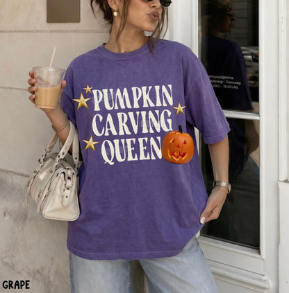 A woman wearing a purple graphic t-shirt with 'PUMPKIN CARVING QUEEN' in white text, gold stars, and a jack-o'-lantern graphic. She is holding a drink and a white handbag, standing in front of a storefront.