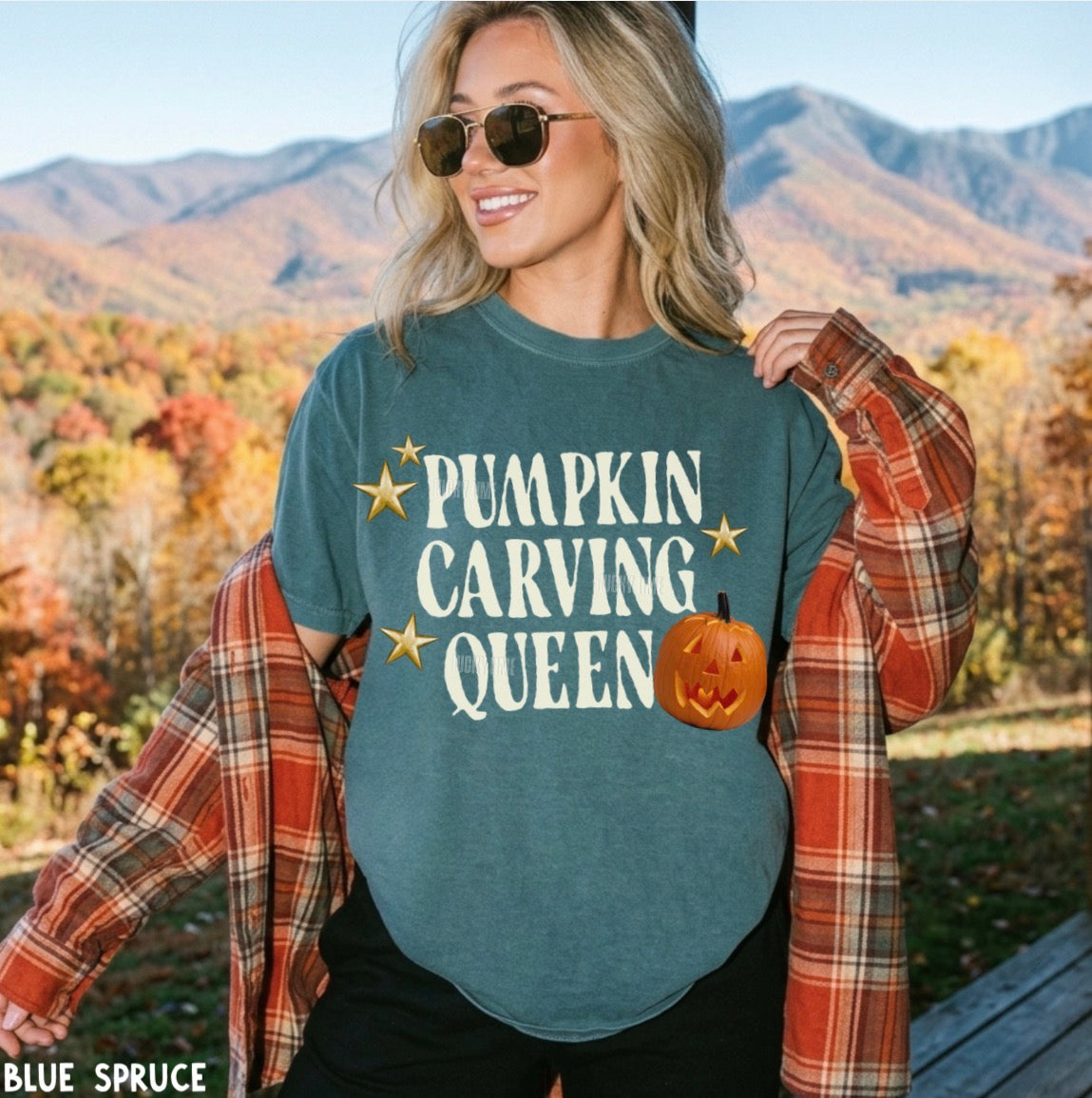 A woman outdoors wearing a slate blue graphic t-shirt that reads 'PUMPKIN CARVING QUEEN' with gold stars and a pumpkin graphic, layered with an orange plaid flannel shirt. The background shows autumn-colored mountains.