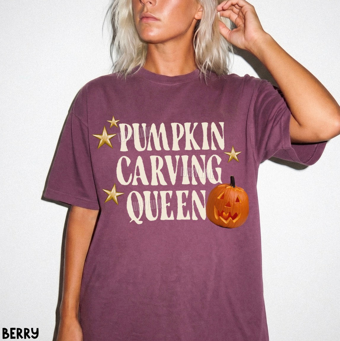 A woman wearing a berry purple t-shirt with a 'Pumpkin Carving Queen' graphic in cream text, gold stars, and an orange jack-o'-lantern, set against a plain white background.