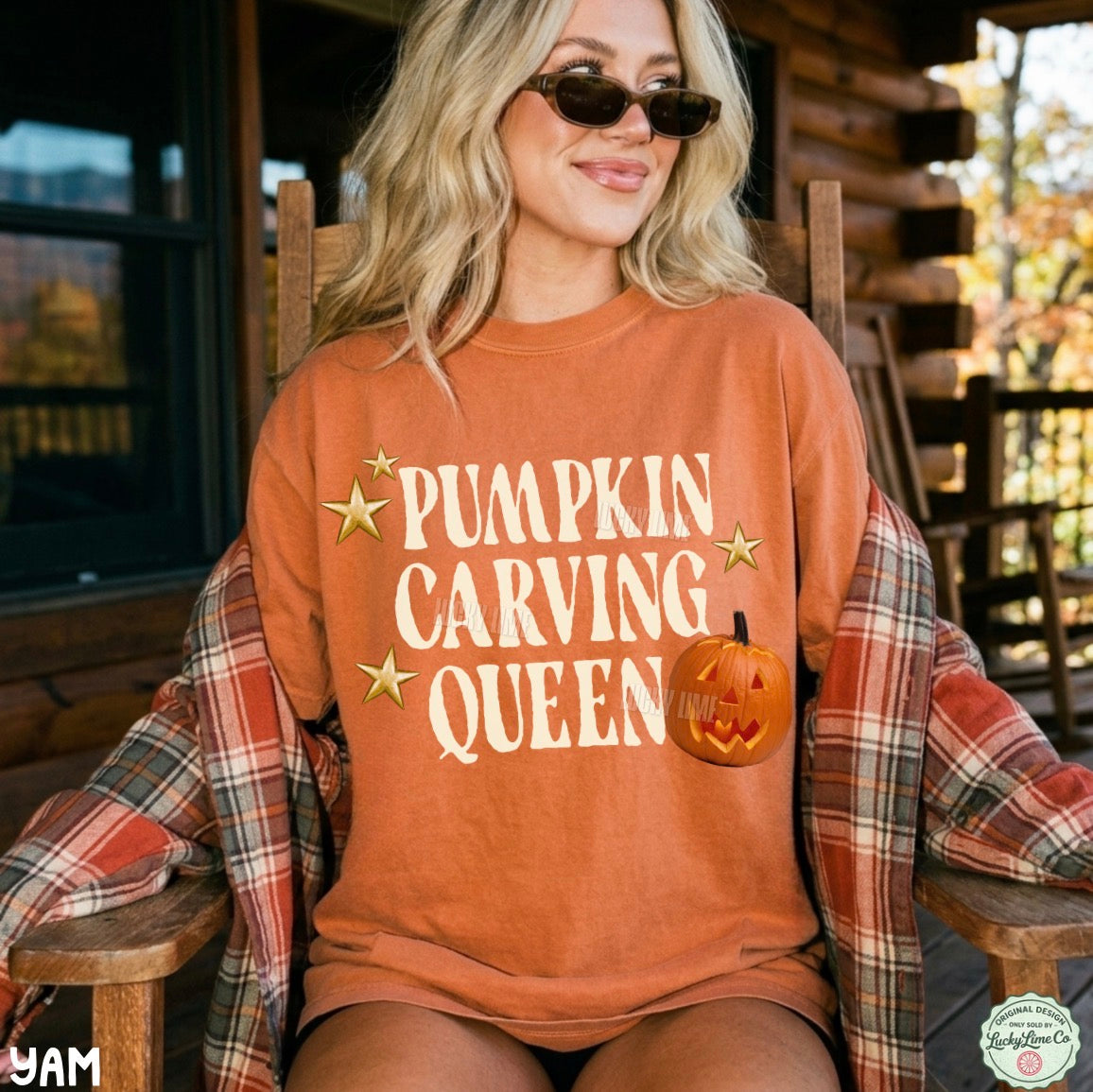 A woman wearing a burnt orange crewneck sweatshirt with 'PUMPKIN CARVING QUEEN' and a jack-o'-lantern graphic, sitting in a wooden rocking chair on a log cabin porch with a plaid shirt draped off her shoulders.