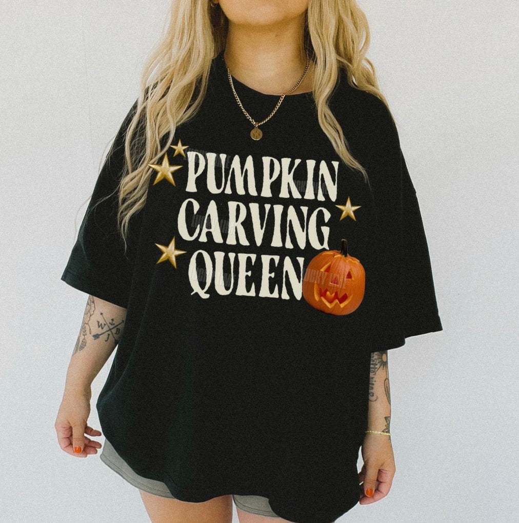 A woman wearing an oversized black graphic t-shirt with the text 'PUMPKIN CARVING QUEEN' in white, gold stars, and a jack-o'-lantern graphic. The image has a white background and a 'BLACK' color label in the bottom left corner.