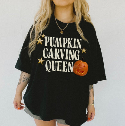 A woman wearing an oversized black graphic t-shirt with the text 'PUMPKIN CARVING QUEEN' in white, gold stars, and a jack-o'-lantern graphic. The image has a white background and a 'BLACK' color label in the bottom left corner.