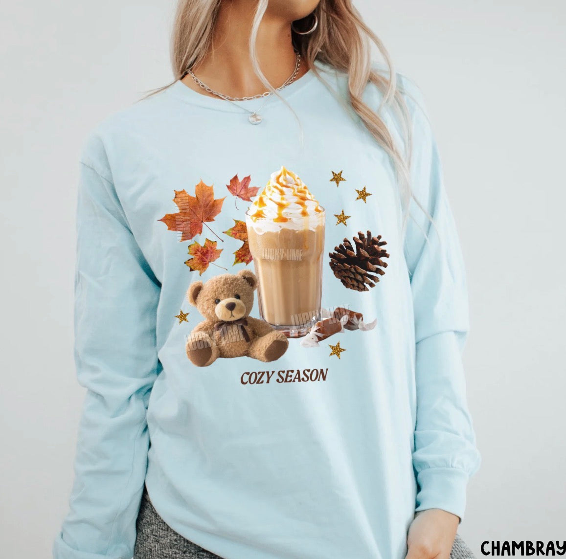 A woman wearing a light blue long-sleeve t-shirt with a cozy autumn graphic featuring a teddy bear, a hot coffee drink, maple leaves, a pinecone, and the text 'COZY SEASON'.