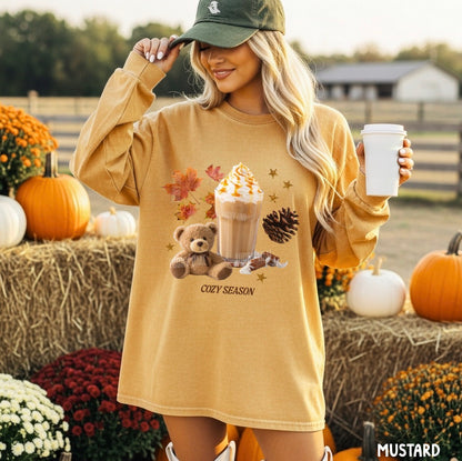 A woman wearing a mustard yellow graphic sweatshirt with a teddy bear and hot chocolate design, posing in an autumn-themed outdoor setting with pumpkins and hay bales.
