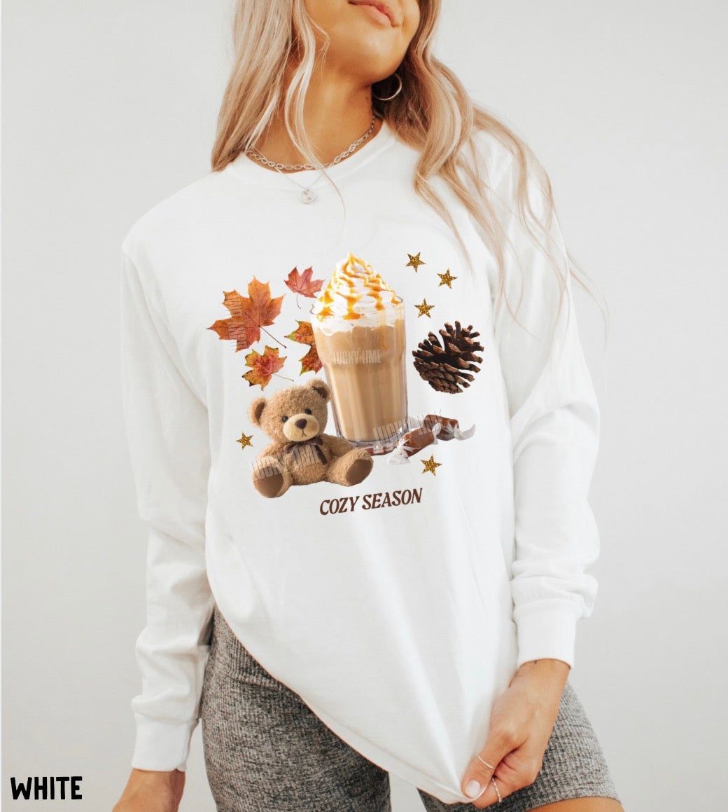 A woman wearing a white long-sleeve sweatshirt with a cozy autumn graphic featuring a teddy bear, a hot coffee drink, maple leaves, pinecones, and the text 'COZY SEASON'.