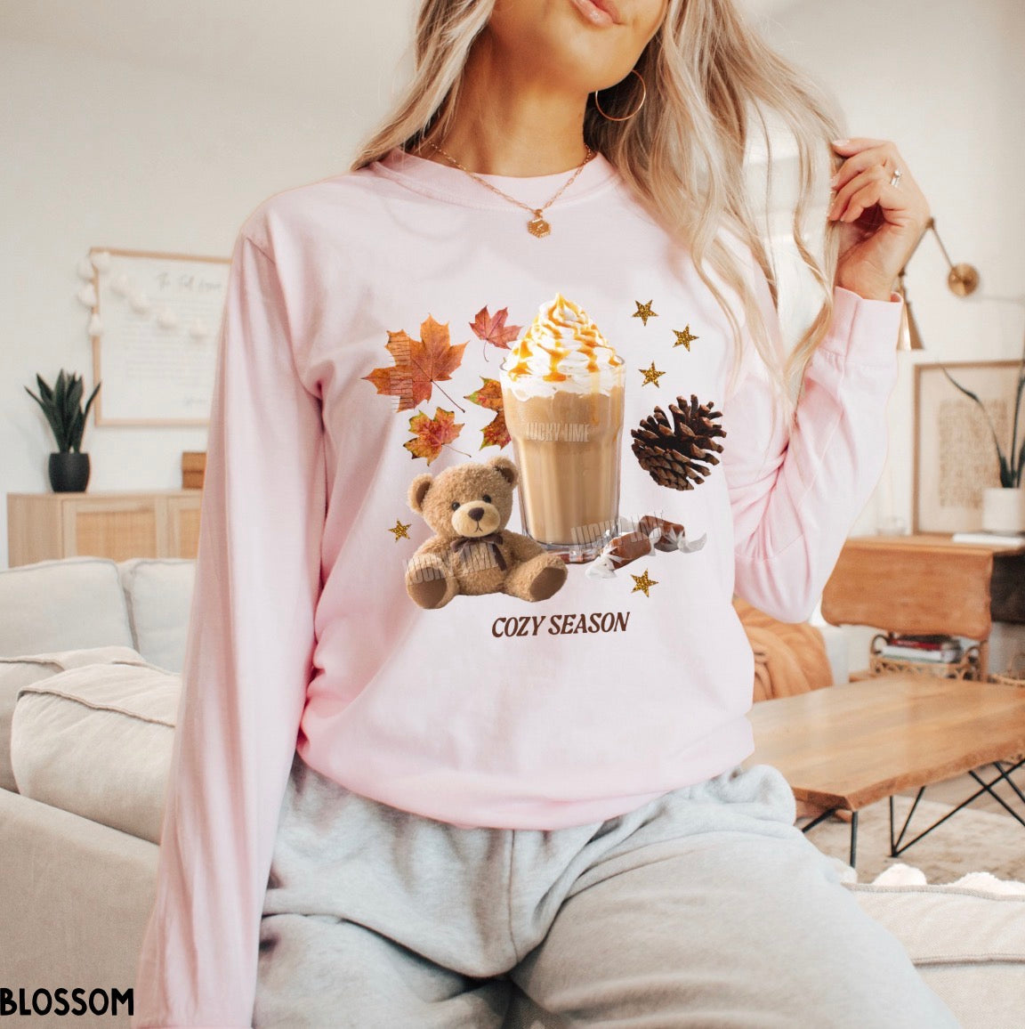 A woman wearing a light pink long-sleeve sweatshirt with a cozy autumn graphic featuring a teddy bear, iced coffee, fall leaves, pinecone, and the text 'COZY SEASON'.