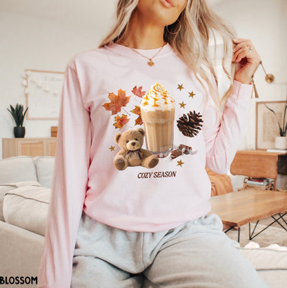 A woman wearing a light pink long-sleeve sweatshirt with a cozy autumn graphic featuring a teddy bear, iced coffee, fall leaves, pinecone, and the text 'COZY SEASON'.