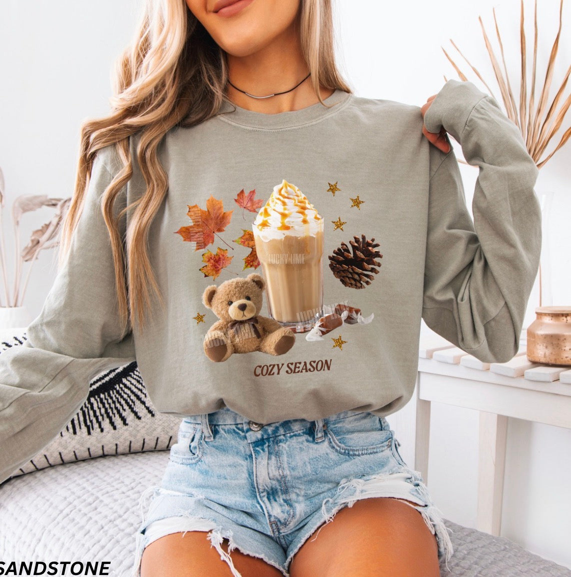 A woman wearing a sage green long-sleeve sweatshirt with a cozy autumn graphic featuring a teddy bear, iced coffee, fall leaves, and pinecones. The text 'COZY SEASON' is printed below the graphic.