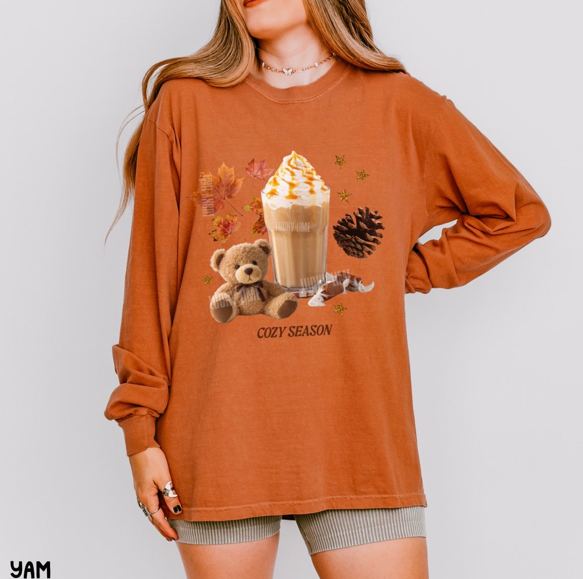 A woman wearing an oversized rust-orange long-sleeve graphic t-shirt with a cozy autumn theme, featuring a hot cocoa drink, teddy bear, pinecone, and maple leaves with the text 'COZY SEASON'.