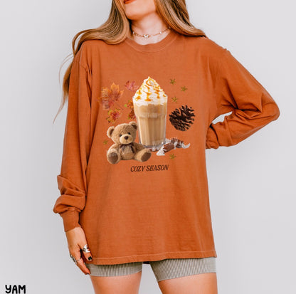 A woman wearing an oversized rust-orange long-sleeve graphic t-shirt with a cozy autumn theme, featuring a hot cocoa drink, teddy bear, pinecone, and maple leaves with the text 'COZY SEASON'.