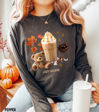 A woman wearing a dark gray long-sleeve graphic sweatshirt with a 'Cozy Season' print featuring a teddy bear, a hot chocolate drink, autumn leaves, and a pinecone. She is holding a white mug and sitting in a cozy indoor setting with pumpkins.