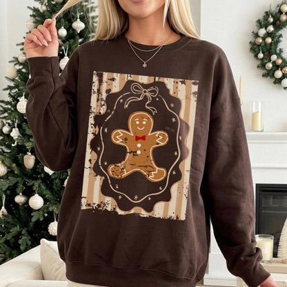 A woman wearing a dark brown crewneck sweatshirt with a large gingerbread man graphic on the front, posed in a cozy living room setting with a decorated Christmas tree and fireplace in the background.