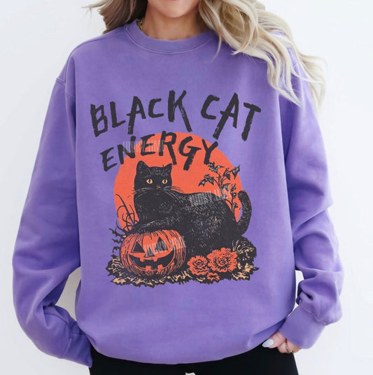 A woman wearing a lavender purple sweatshirt with a Halloween graphic that reads 'BLACK CAT ENERGY' above a black cat, orange pumpkin, and orange sun. The image has a text overlay in the bottom-left corner reading 'VIOLET'.