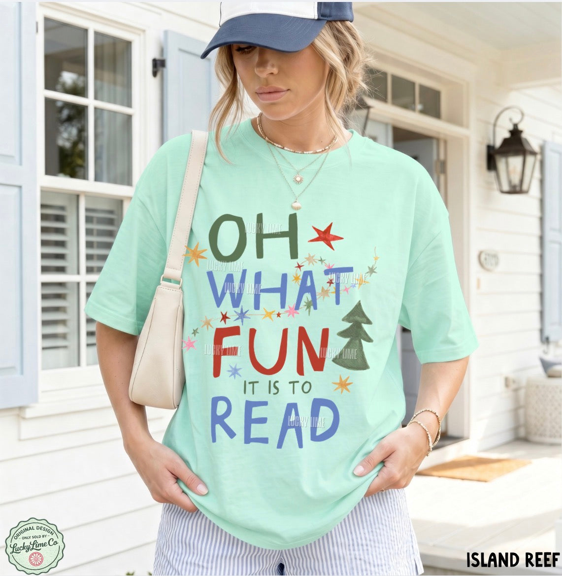 A woman wearing a mint green graphic t-shirt with the text 'OH WHAT FUN IT IS TO READ' in colorful hand-drawn letters, paired with a navy blue visor, striped pants, and a beige shoulder bag, standing in front of a white house porch.