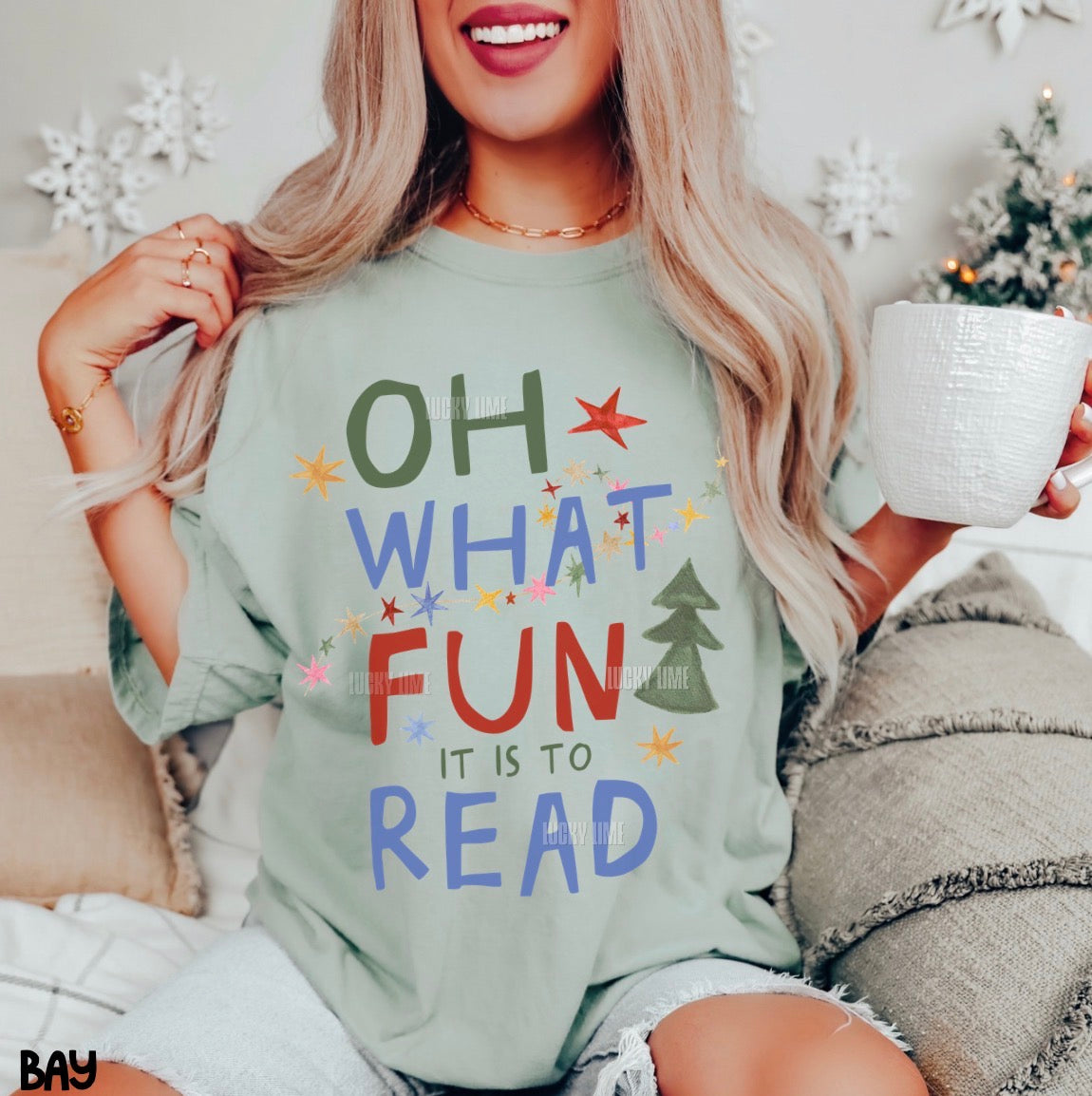 A woman wearing a light sage green graphic t-shirt with the text 'OH WHAT FUN IT IS TO READ' in colorful letters, holding a white mug in a cozy indoor setting.
