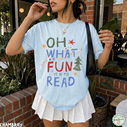 A woman wearing a light blue chambray graphic t-shirt with the text 'OH WHAT FUN IT IS TO READ' in colorful letters, paired with a white pleated skirt, standing in front of a brick wall and potted plants.