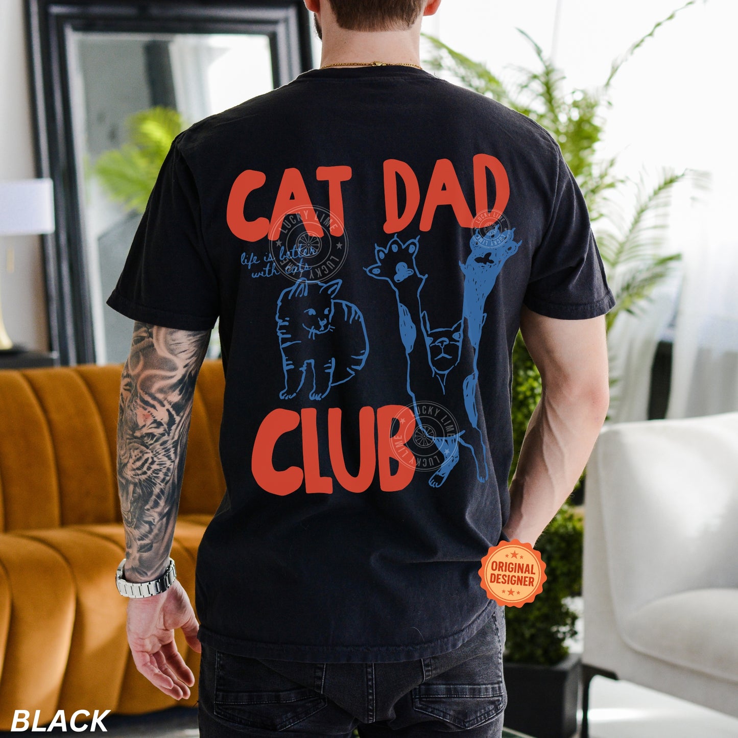 Person wearing a black t-shirt with 'Cat Dad Club' text and cat illustrations in a living room.