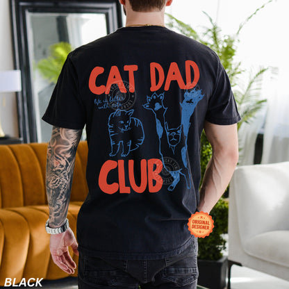Person wearing a black t-shirt with 'Cat Dad Club' text and cat illustrations in a living room.