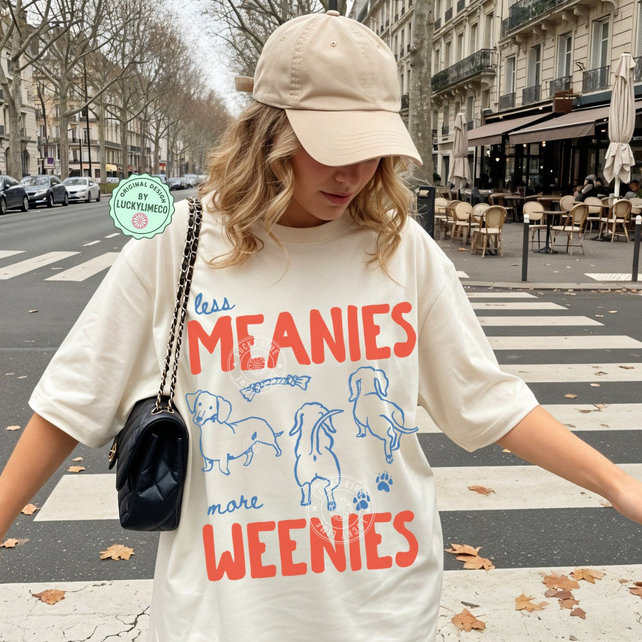 Person wearing an ivory t-shirt with dachshund  graphics and text saying ‘less meanies, more weenies’ standing on a street.