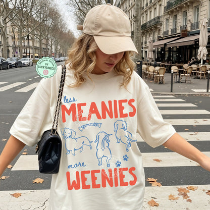 Person wearing an ivory t-shirt with dachshund  graphics and text saying ‘less meanies, more weenies’ standing on a street.
