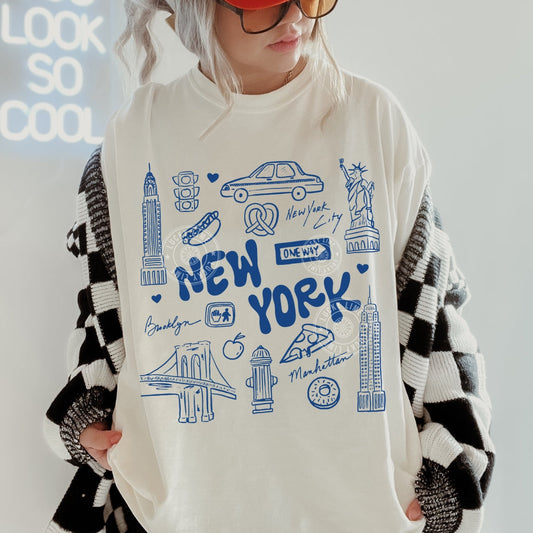 Person wearing a white oversized t-shirt with blue New York-themed graphics and text.